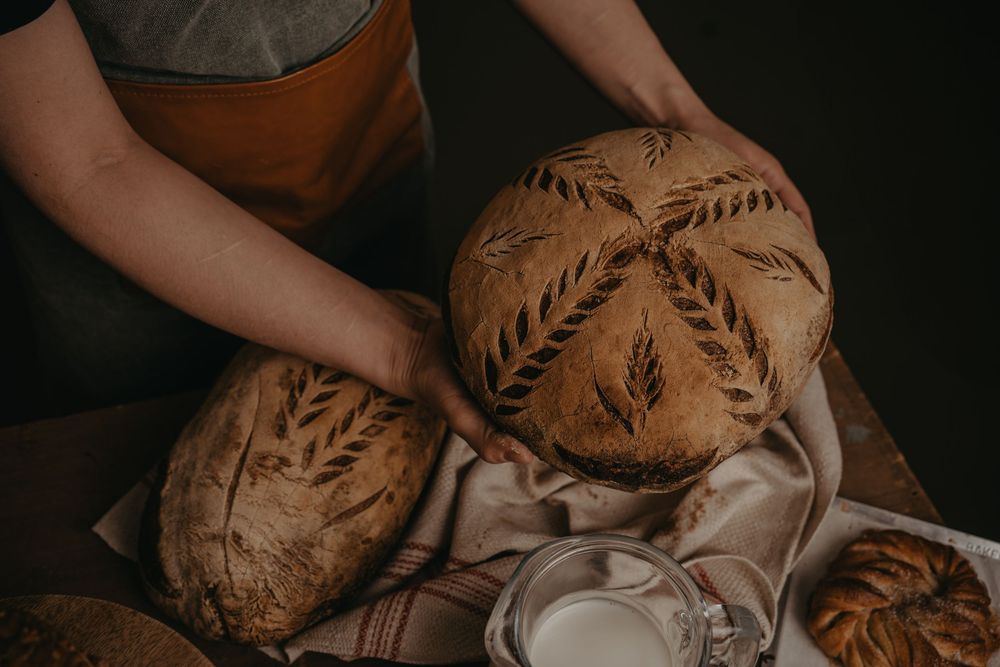 Hands holding freshly baked artisanal sourdough bread with decorative leaf patterns on a linen cloth.