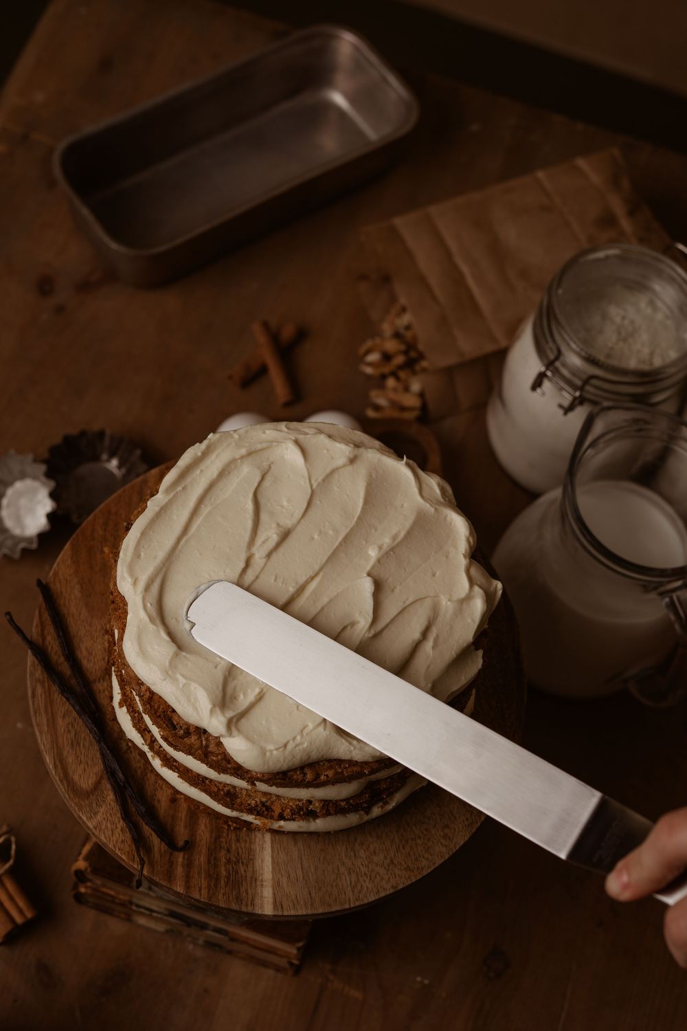 A rustic layered cake being frosted with cream using a metal spatula on a wooden board with mason jars nearby.