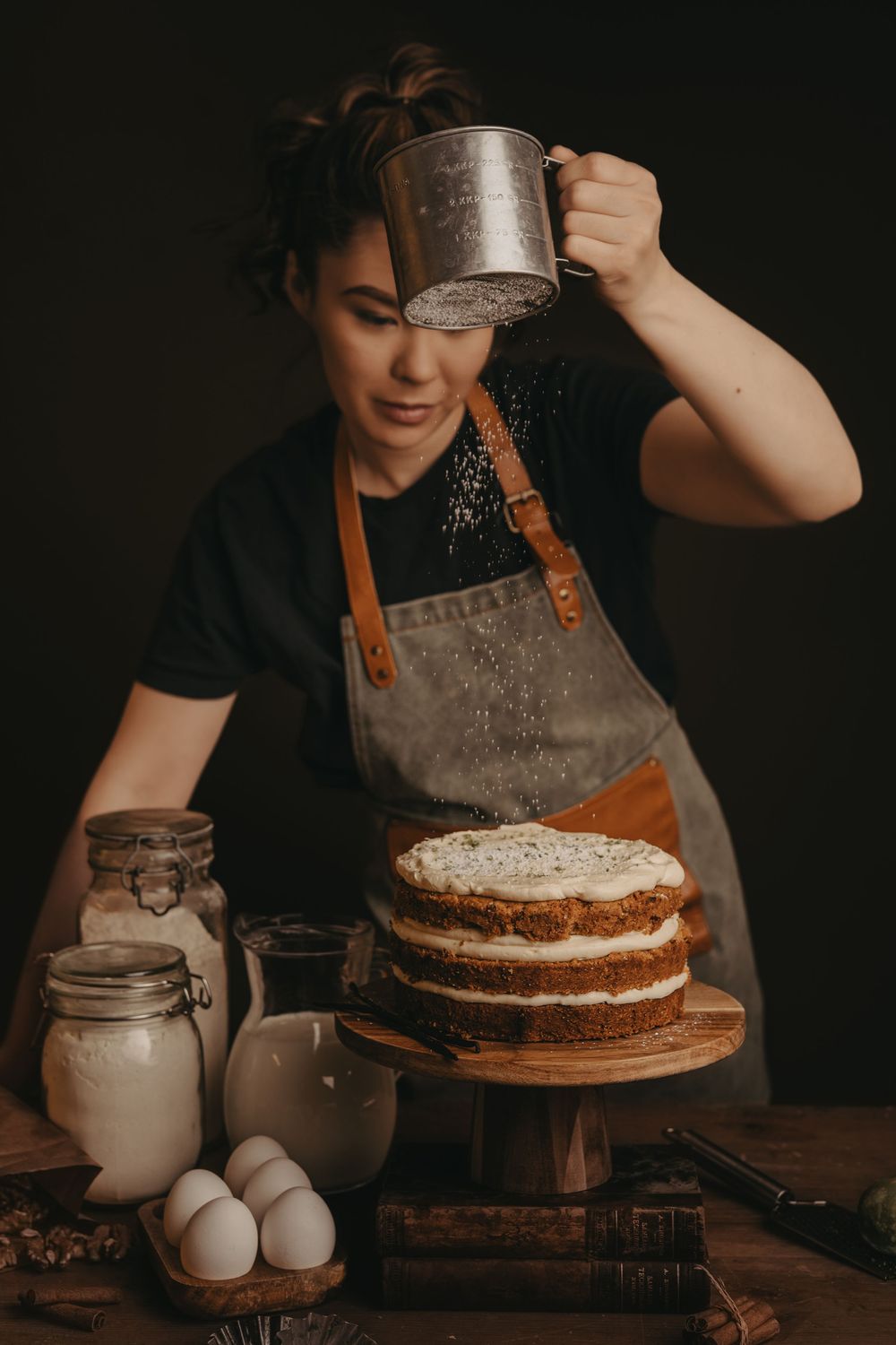 Dark moody baking scene showing preparation of a layered cake with cream filling and rustic jars of ingredients on wooden surface.