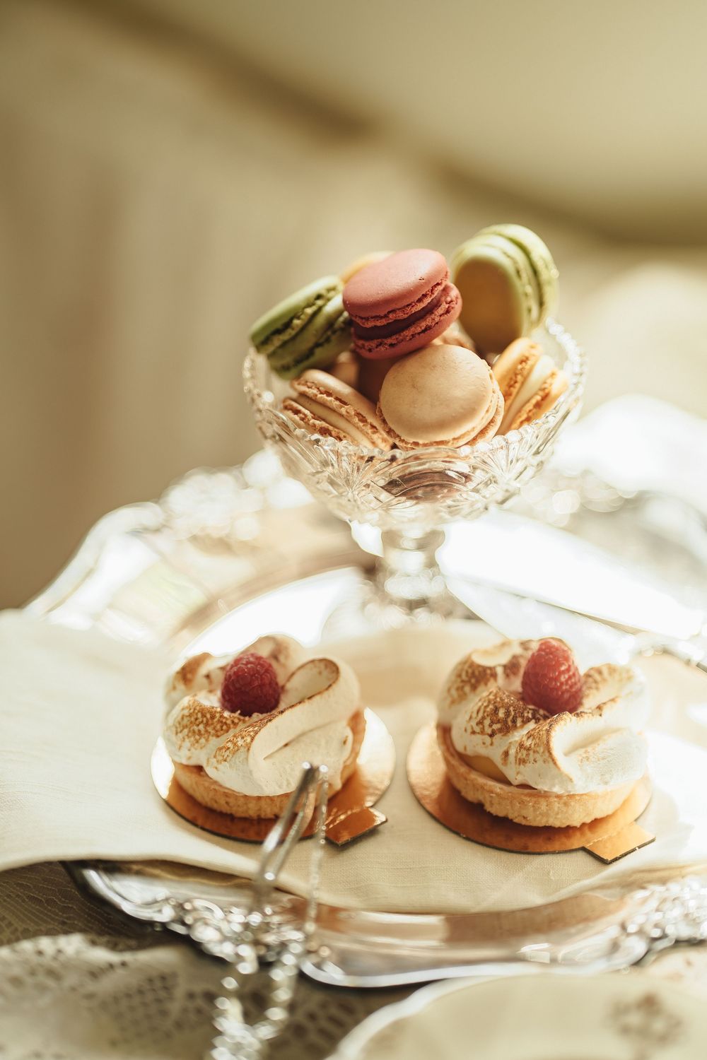 Elegant dessert display with macarons in crystal bowl and raspberry meringue tarts on silver tray.