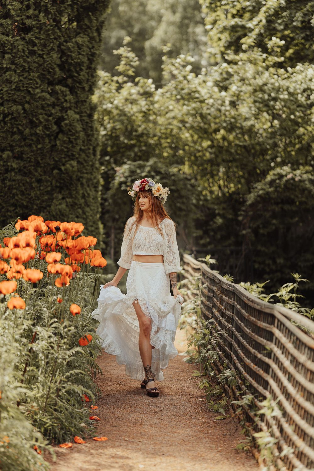Woman in floral crown and white boho dress walks along garden path lined with vibrant orange poppies and lush greenery.