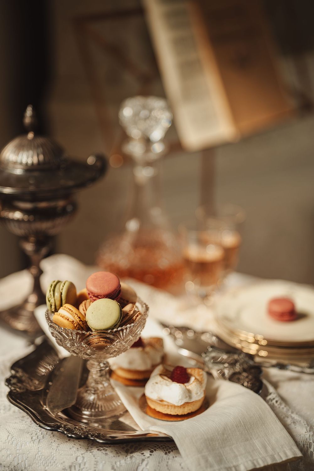 Elegant tea party display with colorful macarons in crystal bowl, cream pastries, and vintage silver serving pieces.