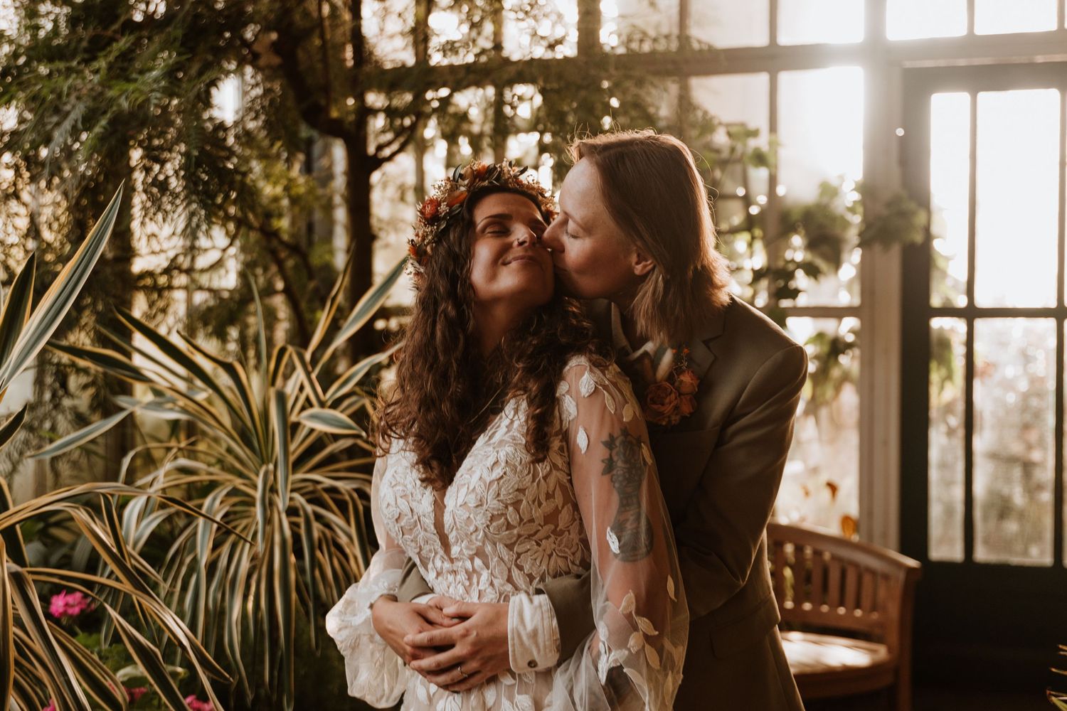 A romantic couple embraces in a sunlit greenhouse surrounded by lush plants and natural light.