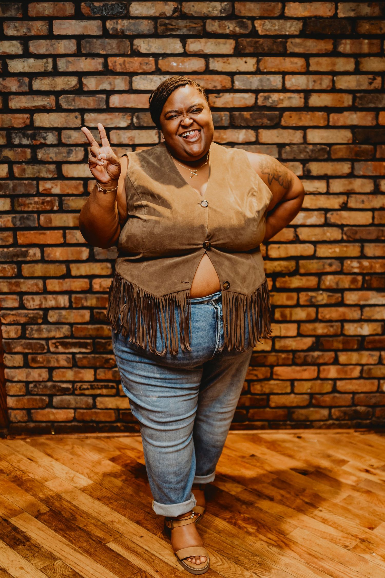Person wearing fringed brown top and jeans posing against brick wall.