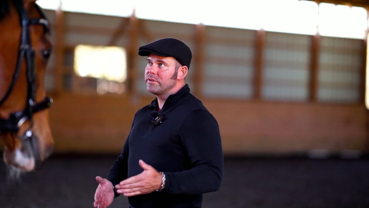 A person in a black quarter-zip and baseball cap gestures while speaking inside a horse stable.