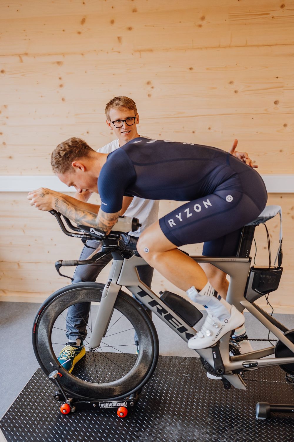 Cyclist in aerodynamic position training on an indoor stationary bike against wooden wall background.