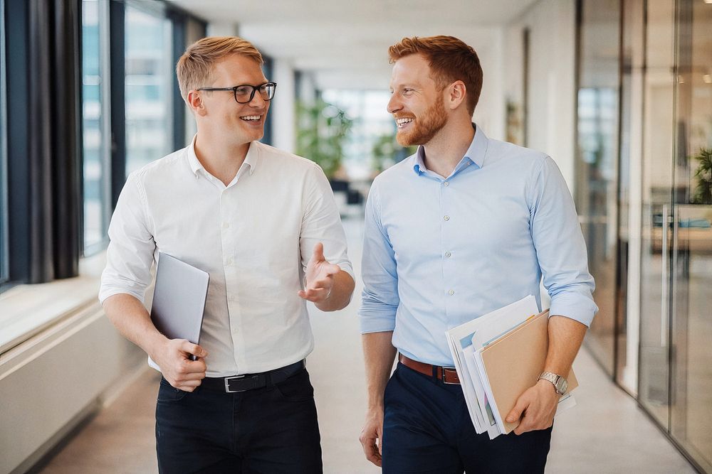 Two colleagues in business attire walking and having an animated conversation in a bright modern office hallway.