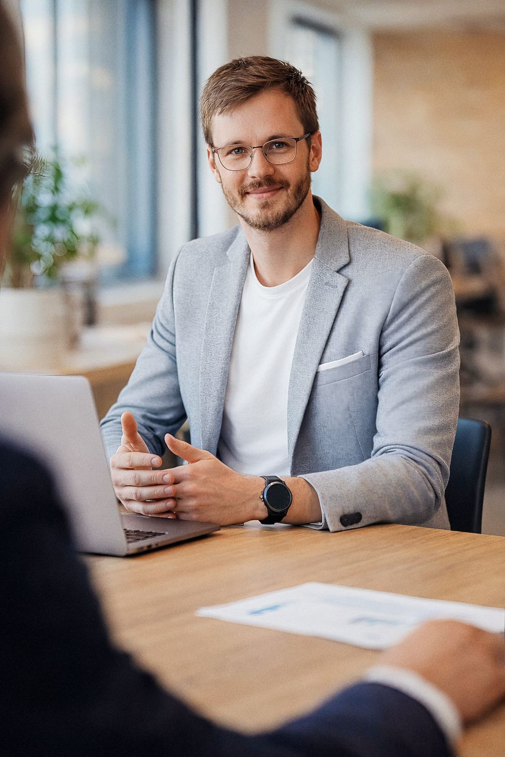 Professional in a gray blazer and white shirt sits at desk during business meeting with laptop and smartphone.