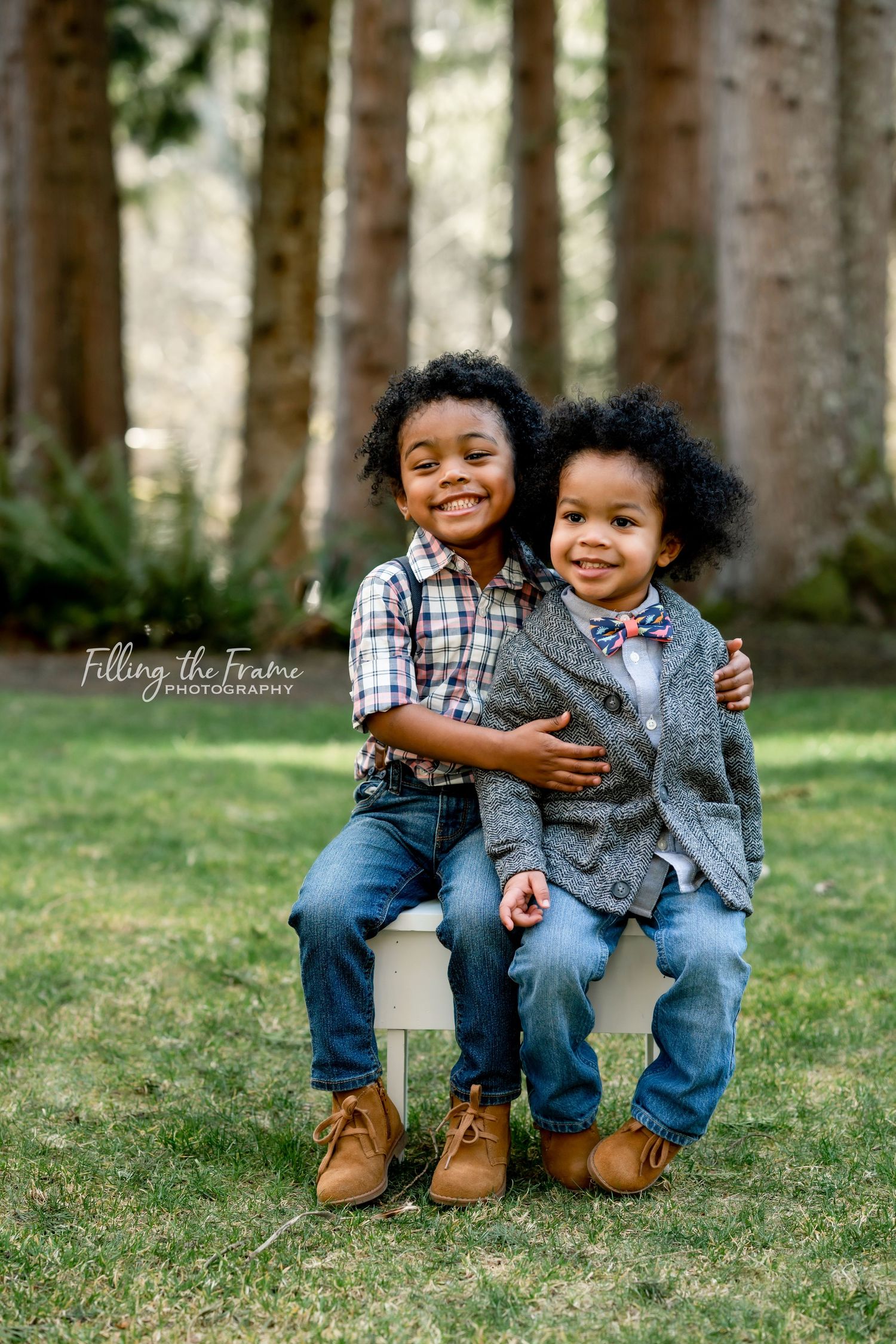 Two young children sit together on a wooden bench outdoors sharing a sweet embrace in matching denim outfits.