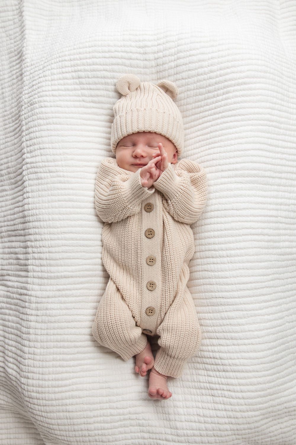 A newborn baby wearing a cozy cream-colored knit sweater lies on soft white textured bedding.