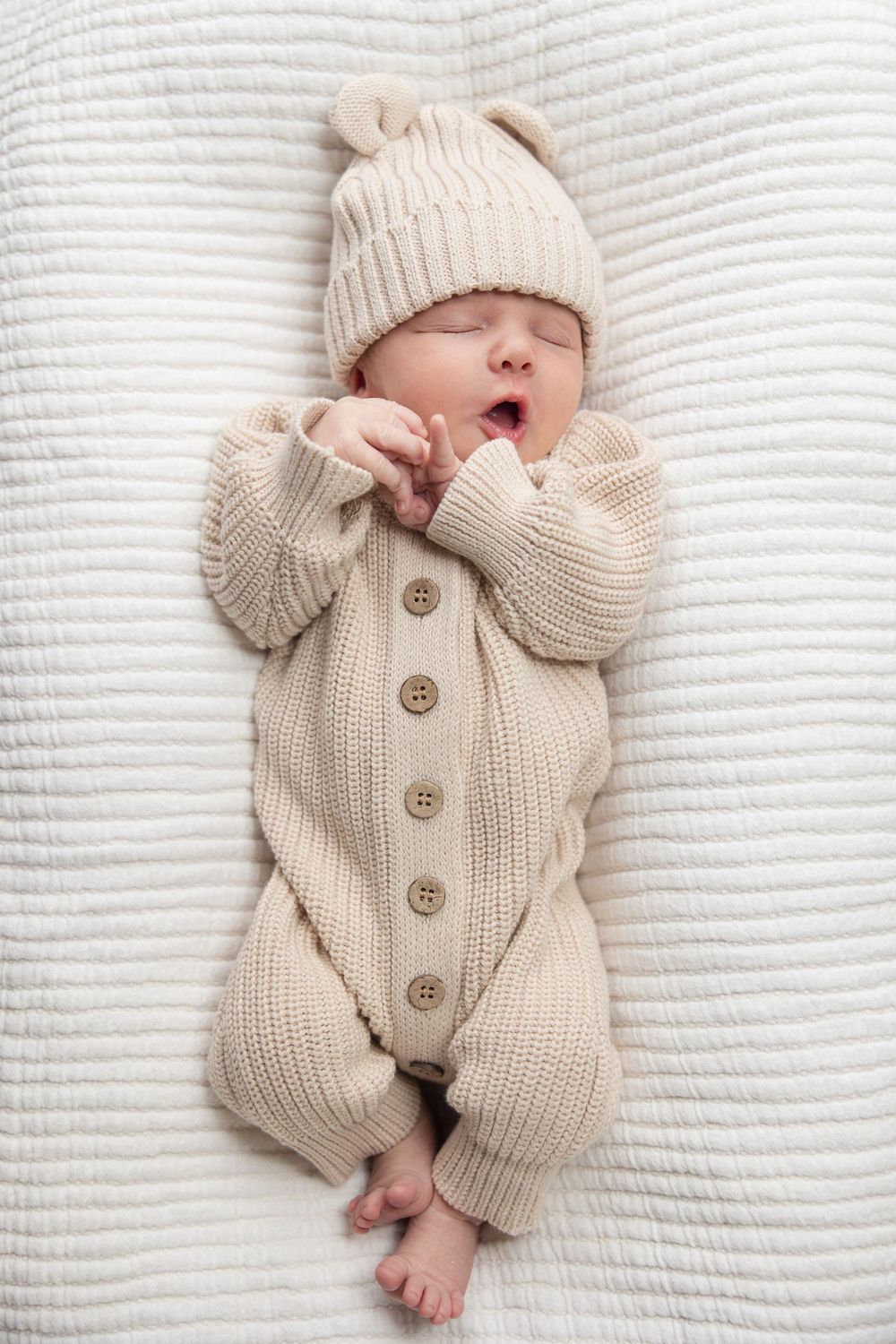Sleeping baby wearing cozy cream knit romper and matching hat with ears on white textured blanket.