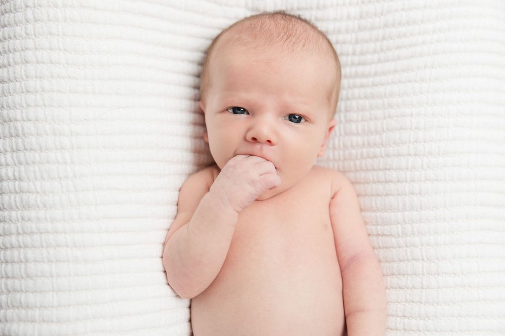 A small baby lies on white textured fabric while sucking their thumb.