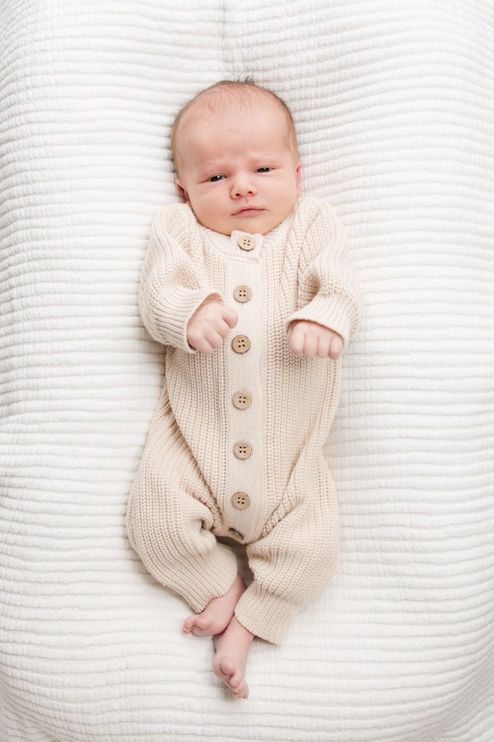 A small baby wearing a cozy cream-colored knit romper with wooden buttons lies peacefully on white textured bedding.