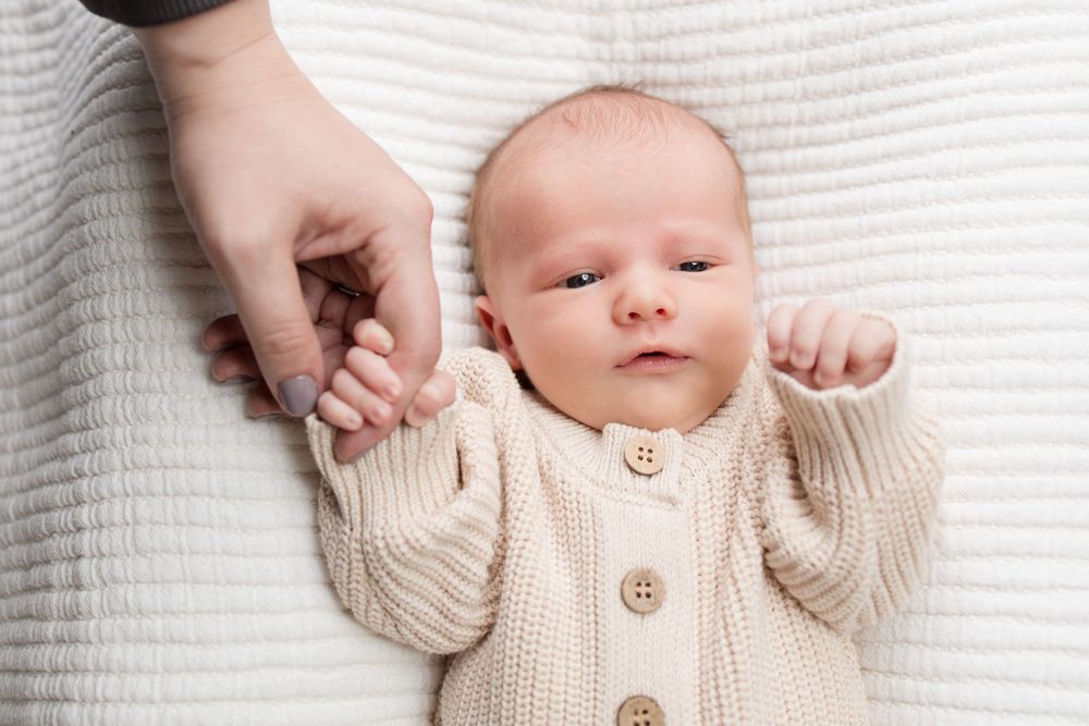 A newborn baby wearing a cream knit cardigan lies on white fabric while someone gently holds their tiny hand.