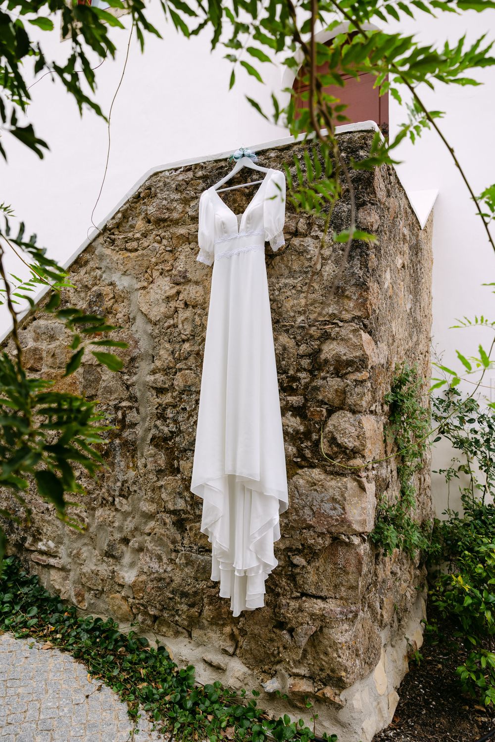 White wedding dress hanging on stone wall with green foliage in outdoor garden setting in Mafra near Lisbon.
