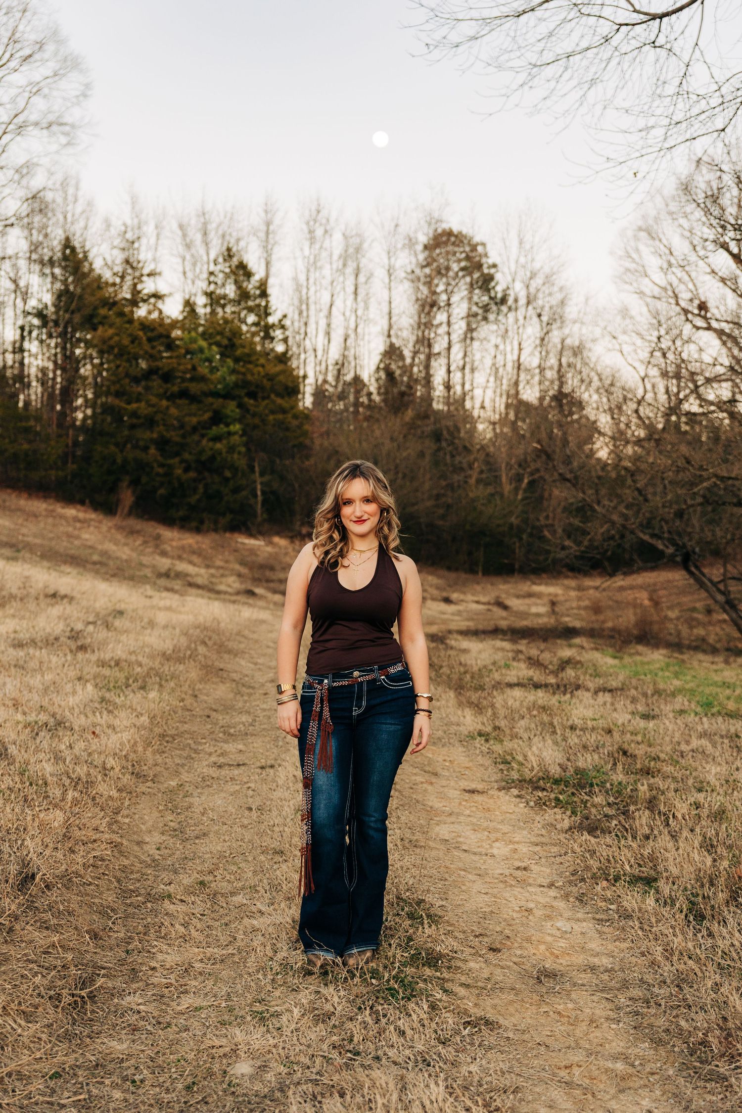 Someone in a dark tank top and flared jeans stands on a dirt path surrounded by bare trees at sunset.