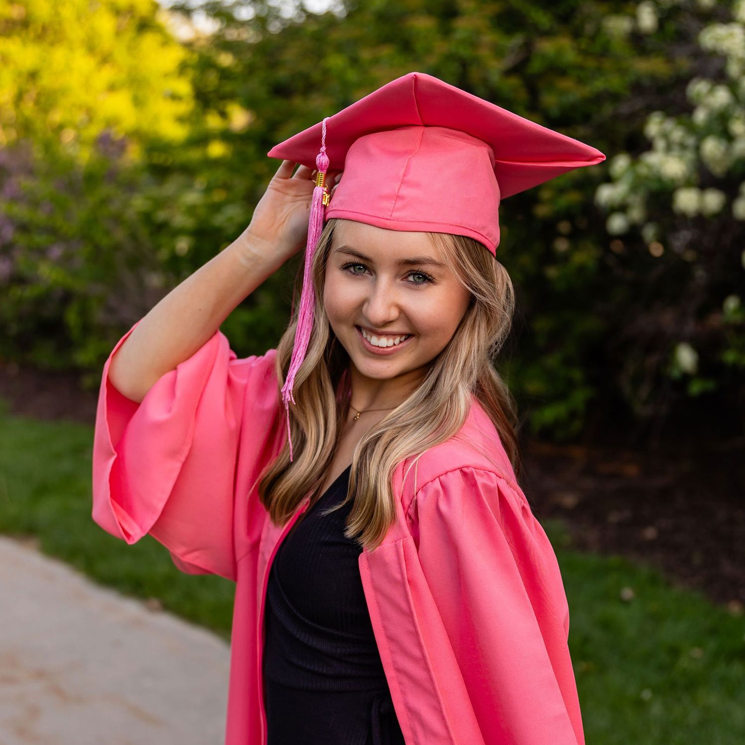 Graduate i pink cap and gow outdoors against green foliage during spring graduation photoshoot at OPPD Arboretum in Omaha, NE.