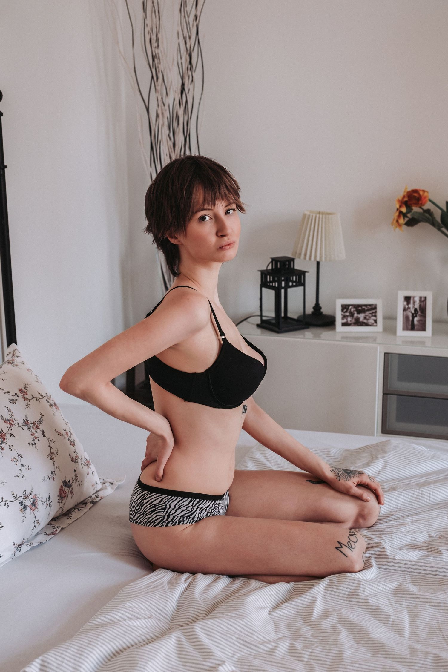 Person in black sports bra and patterned shorts poses on white bedding in a cozy bedroom setting with decor.