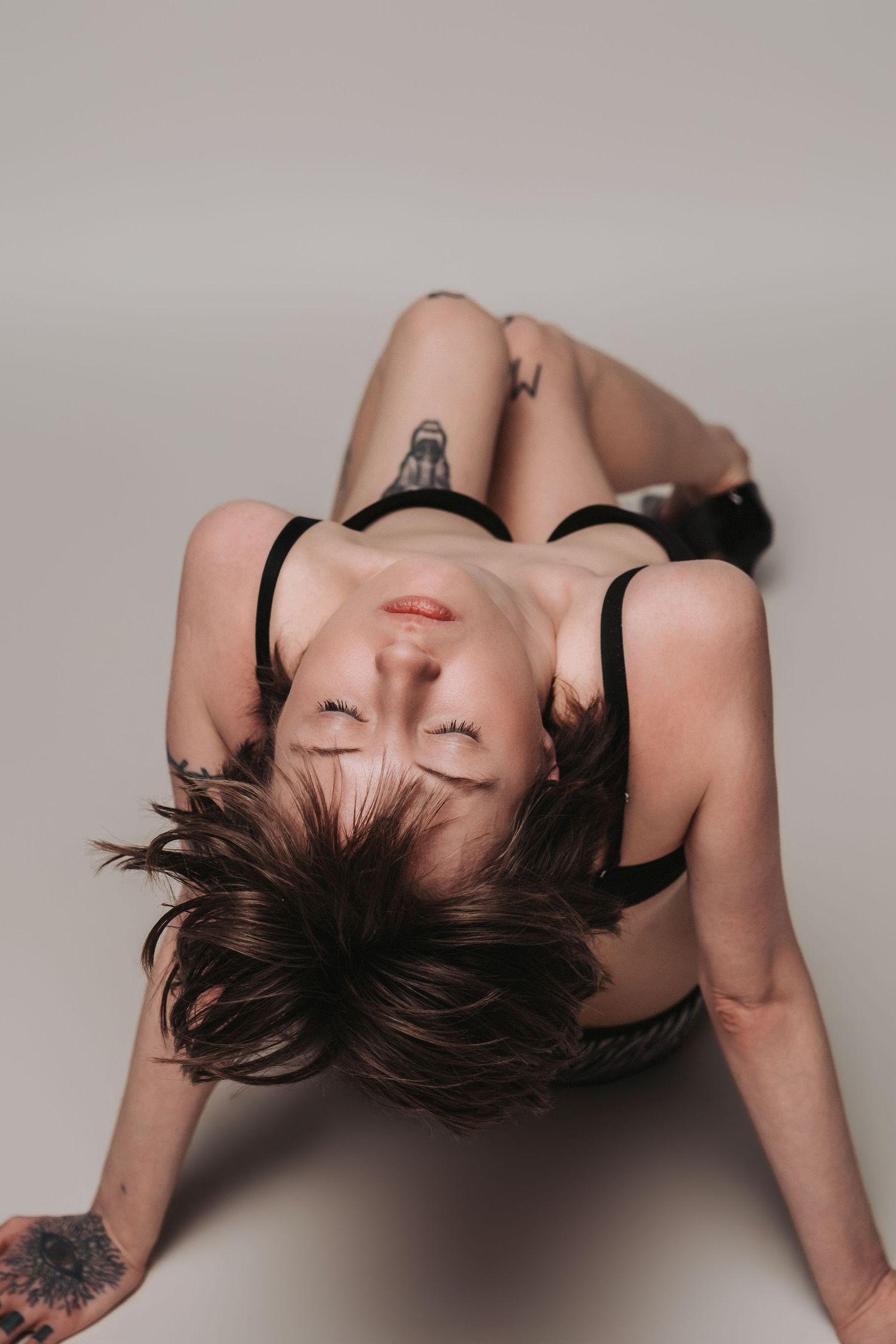 Artistic boudoir portrait of person with dark hair lying back in black lingerie against neutral background.