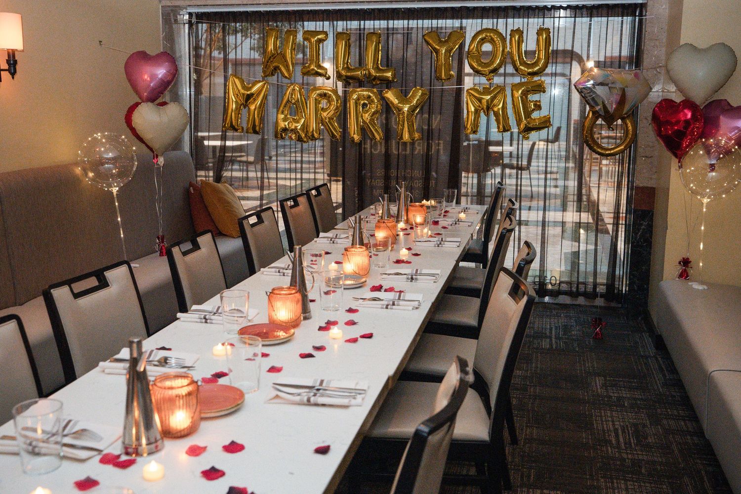Romantic dinner setup with gold 'MARRY ME' balloon letters, white tablecloth, and heart decorations for a proposal.