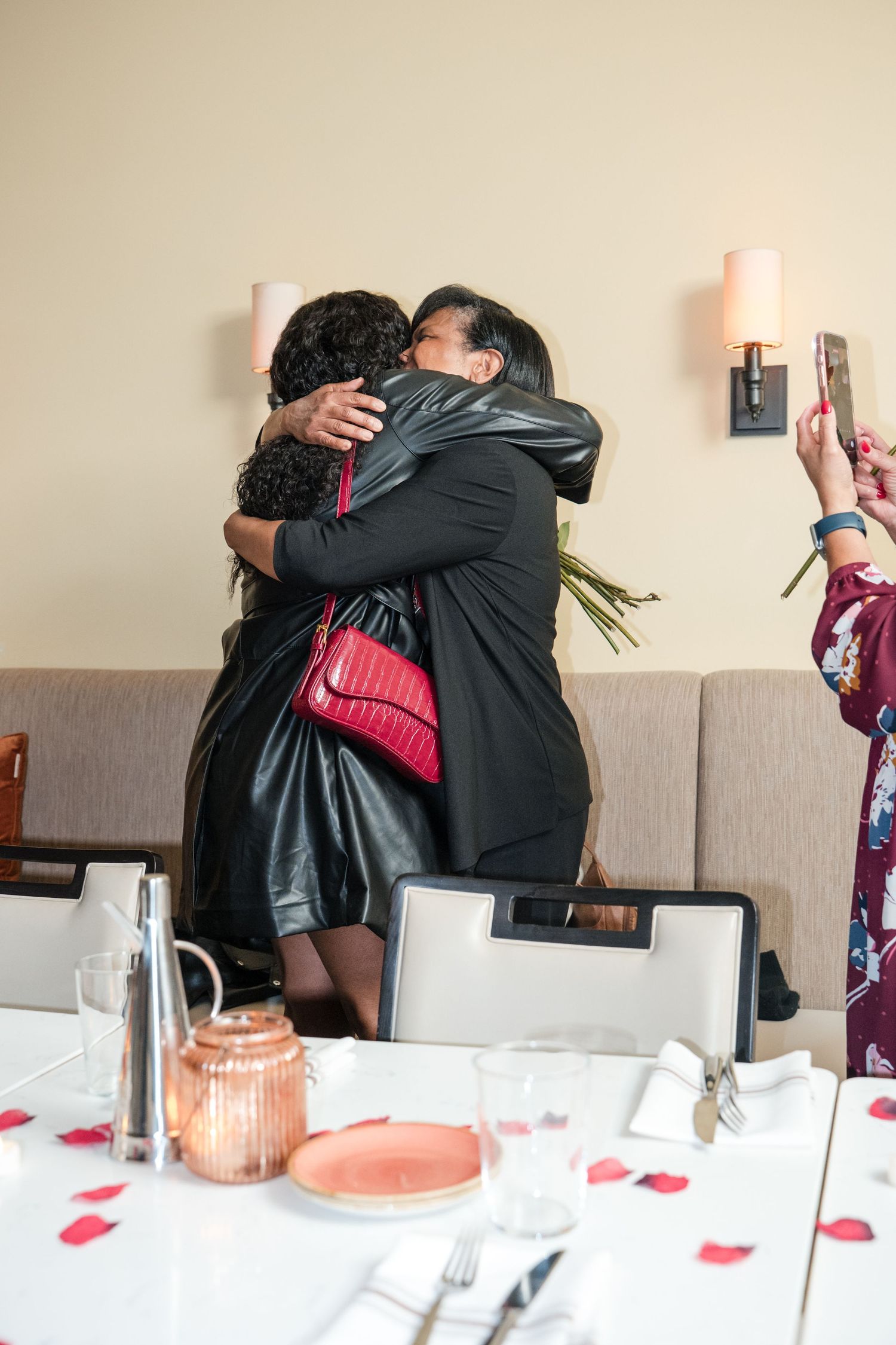 Two people share an emotional embrace at a celebration with rose petals scattered on a dining table.