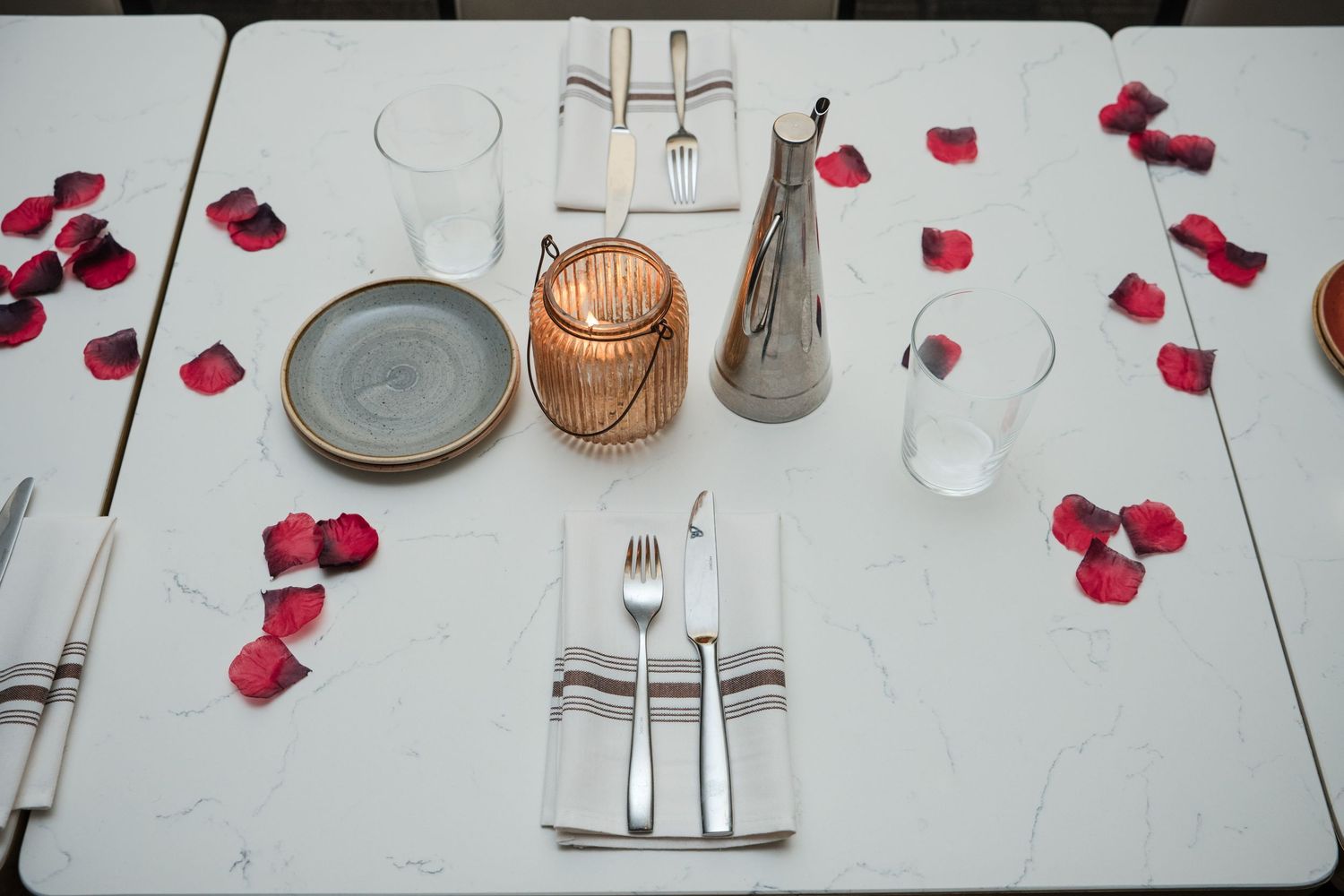 Elegant table setting with scattered red rose petals and glowing candles on white tablecloth.