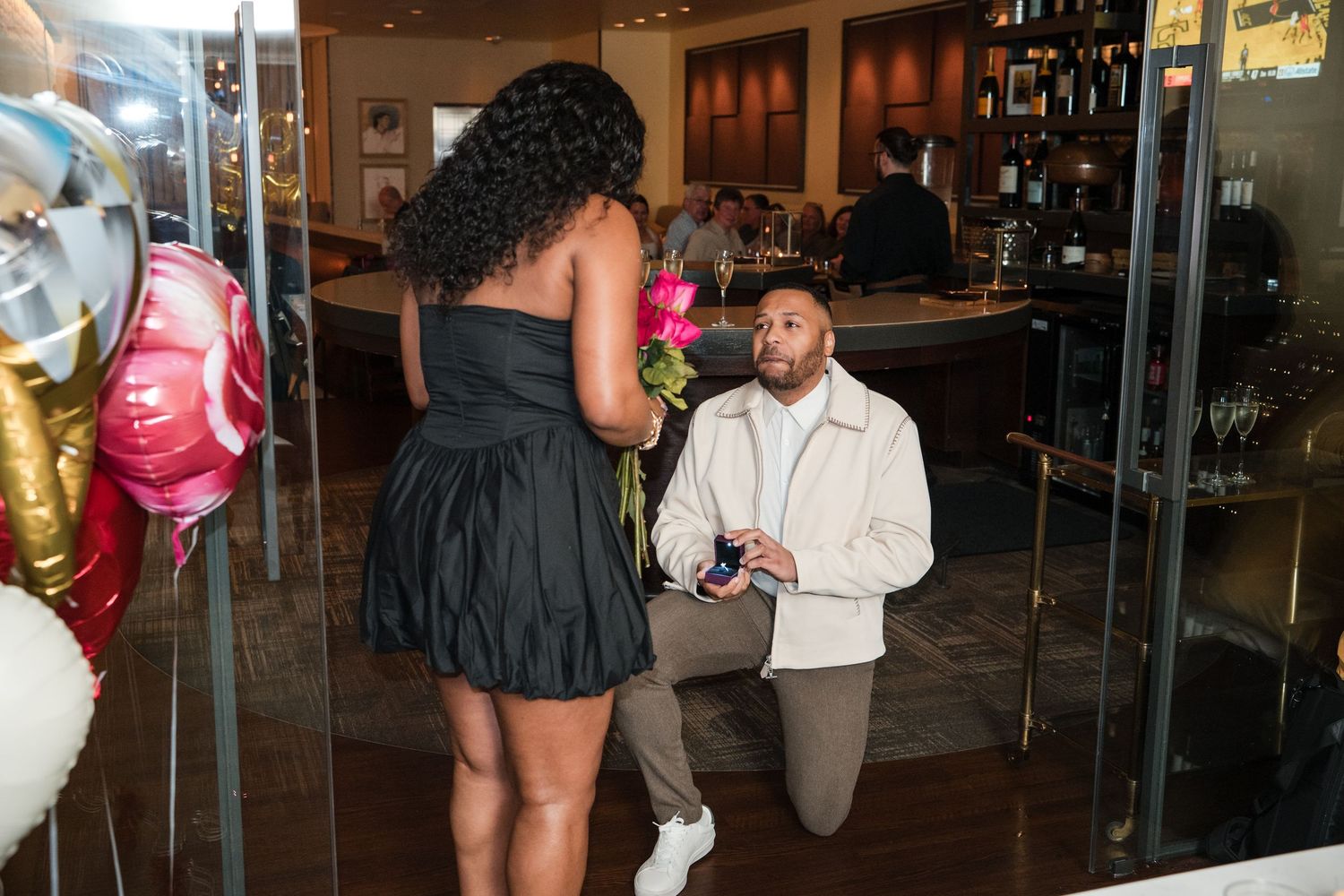 A couple shares an intimate moment during a proposal at an indoor event venue with guests looking on.