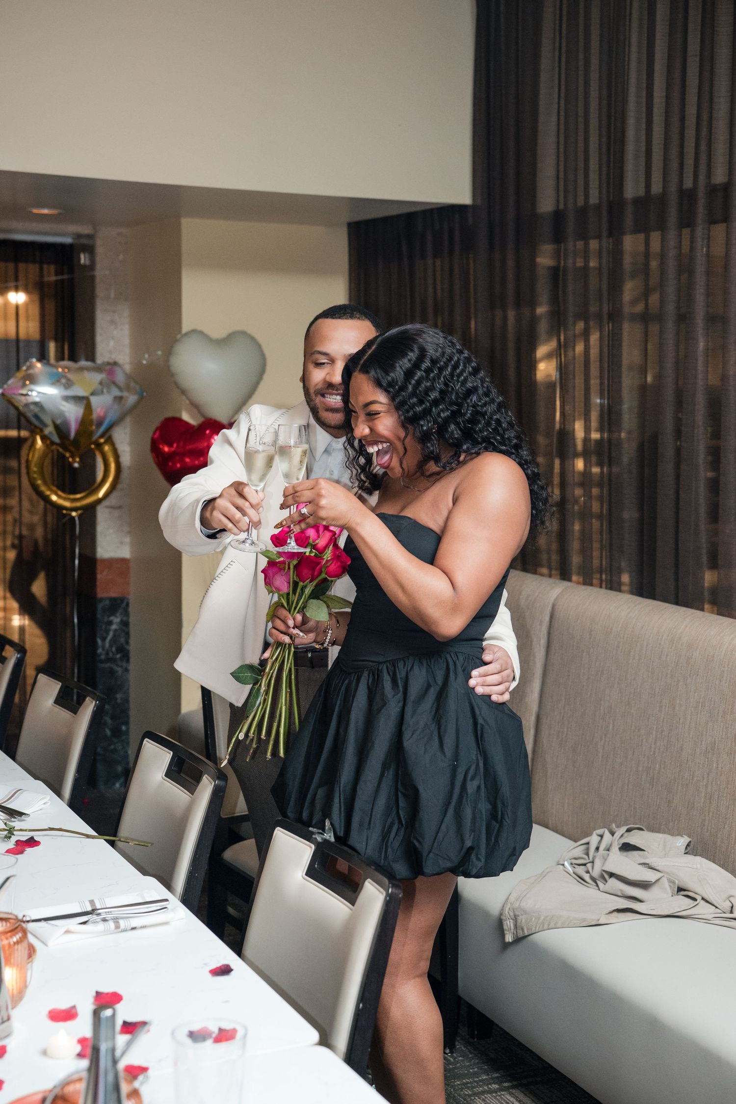 A couple celebrates with champagne and flowers at an intimate gathering with heart-shaped balloons in the background.