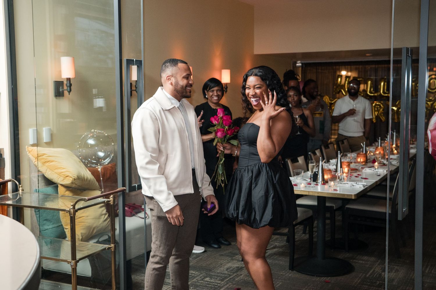 An indoor proposal celebration with guests watching through glass partitions.