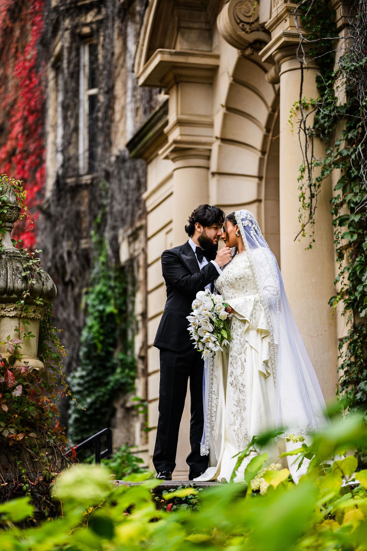 A romantic wedding couple embraces near a stone building with vibrant red ivy and lush greenery.