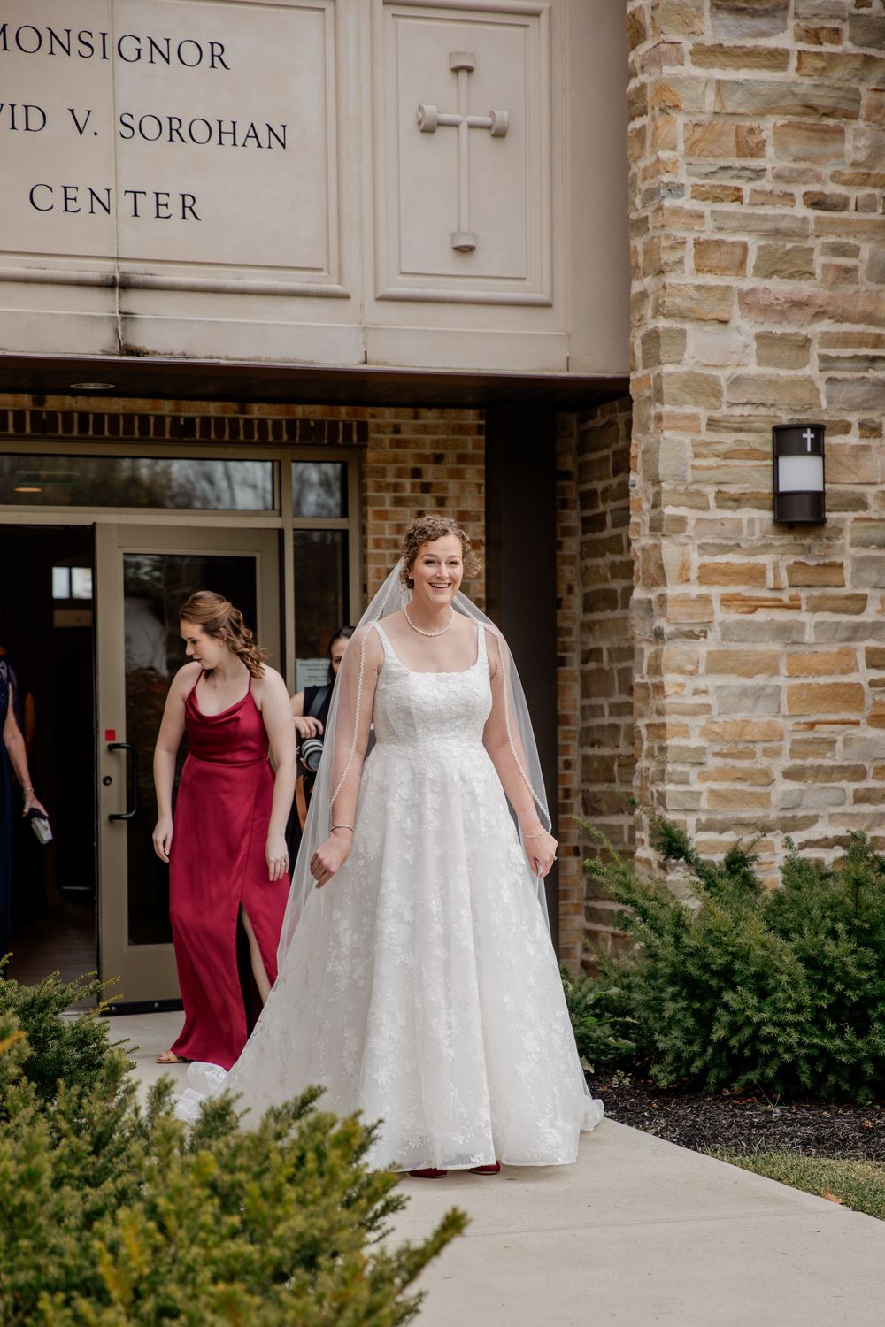 A wedding party member in a burgundy dress accompanies a bride in a white lace gown outside a brick church entrance.