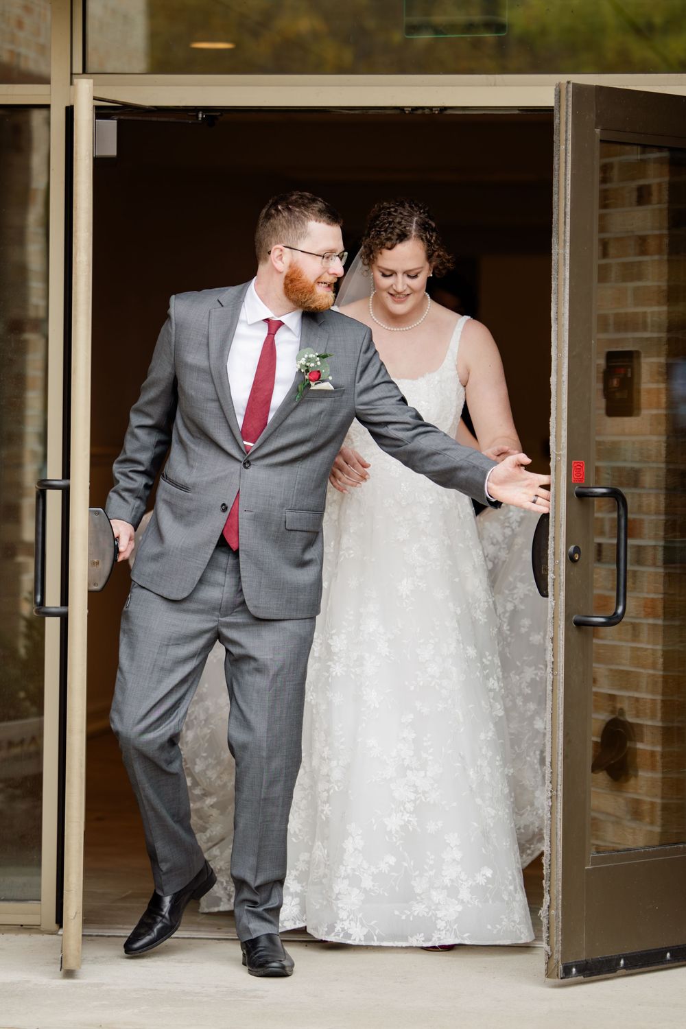 Wedding couple shares a joyful moment while walking through glass doors in matching gray and white attire.