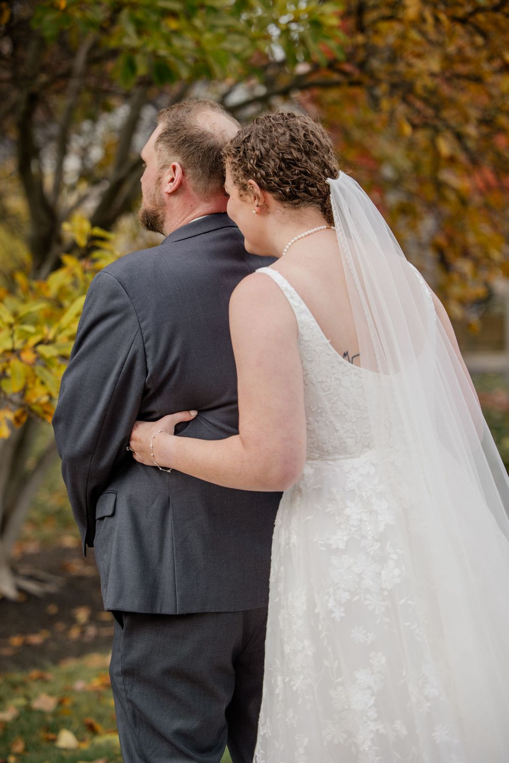 A bride in a white dress and veil embraces her companion wearing a dark suit during an autumn wedding photoshoot.