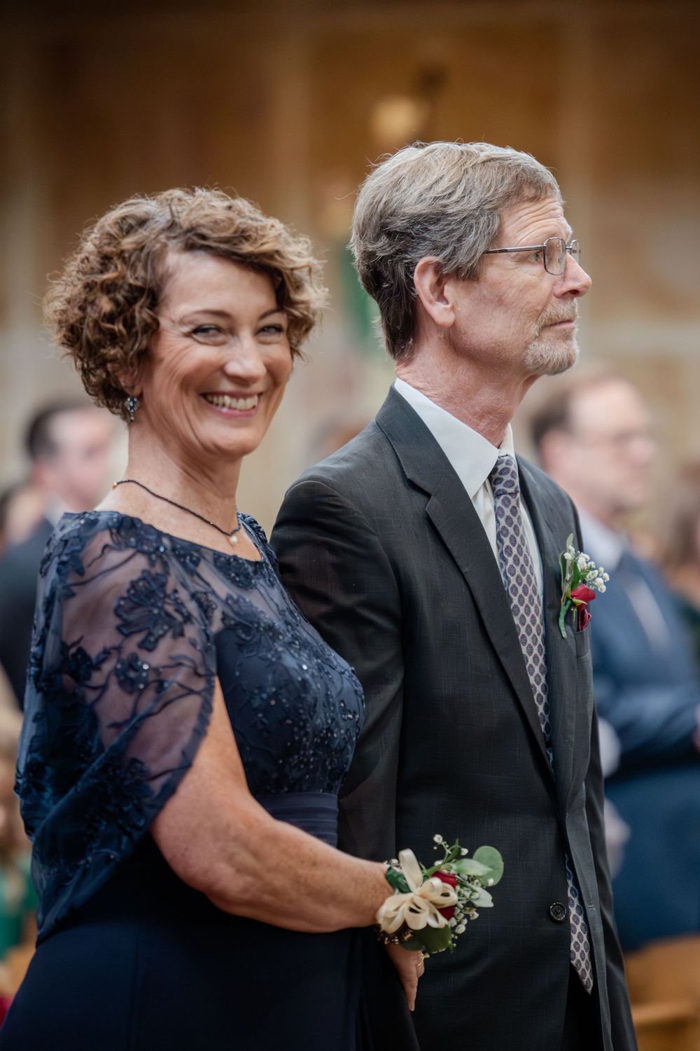 A couple in formal attire at a wedding ceremony, with the woman wearing a navy lace dress and the man in a dark suit.