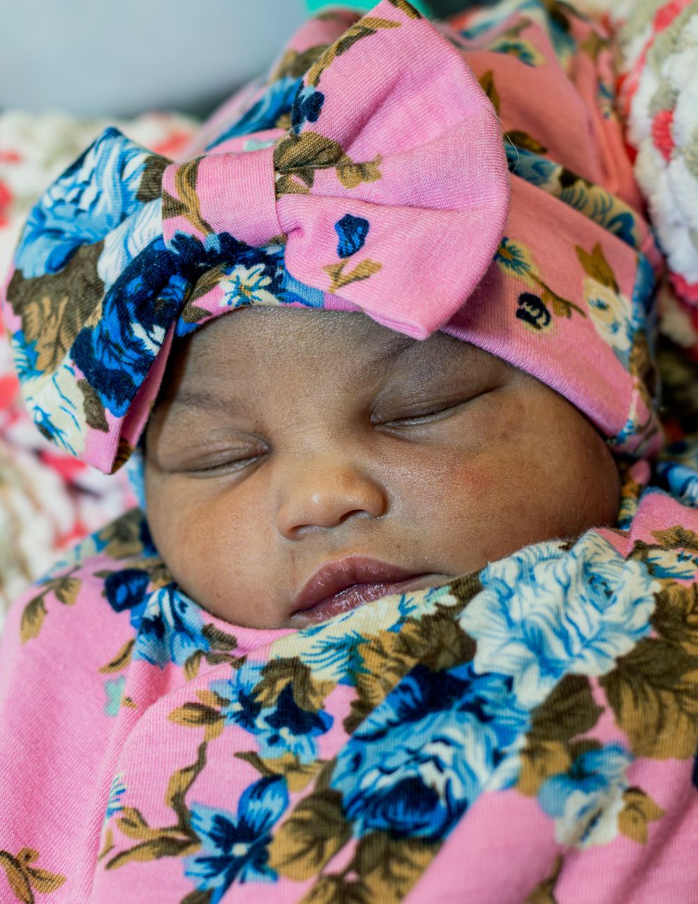 A sleeping newborn wearing a pink and blue floral headband against matching bedding.