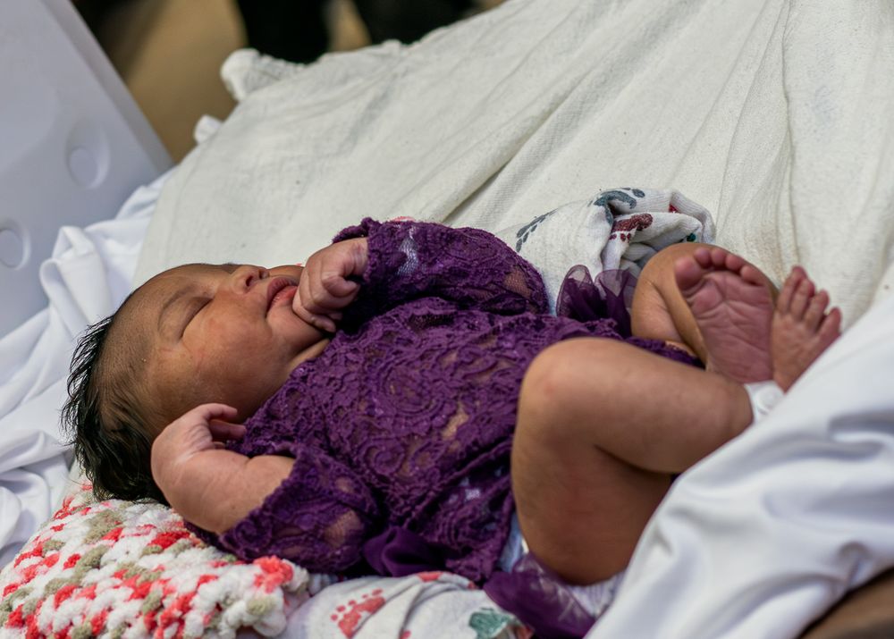 A newborn baby wearing purple lace clothing lies on floral bedding.
