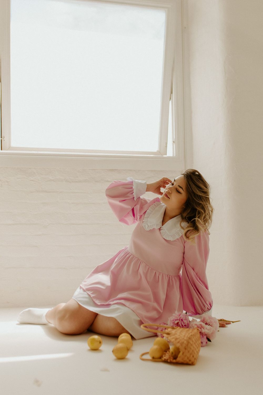 A person in a pink dress lies on a white floor surrounded by citrus fruits in a bright sunlit room.