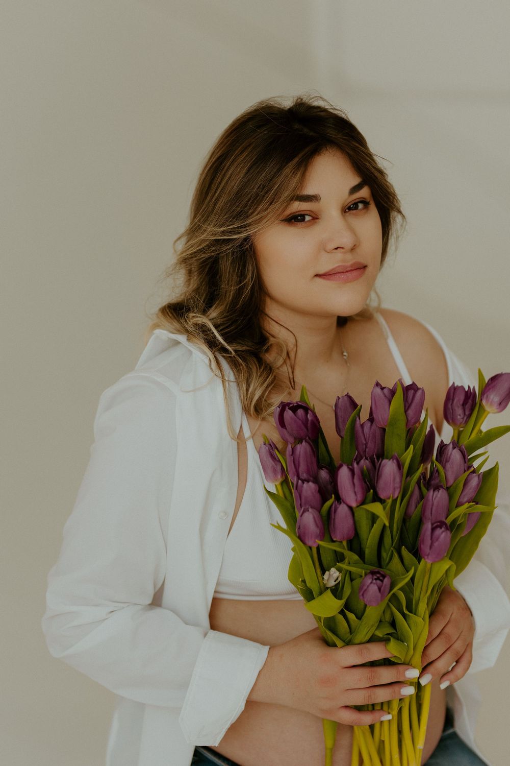 A woman in a white blouse poses with a bouquet of purple tulips against a neutral background.