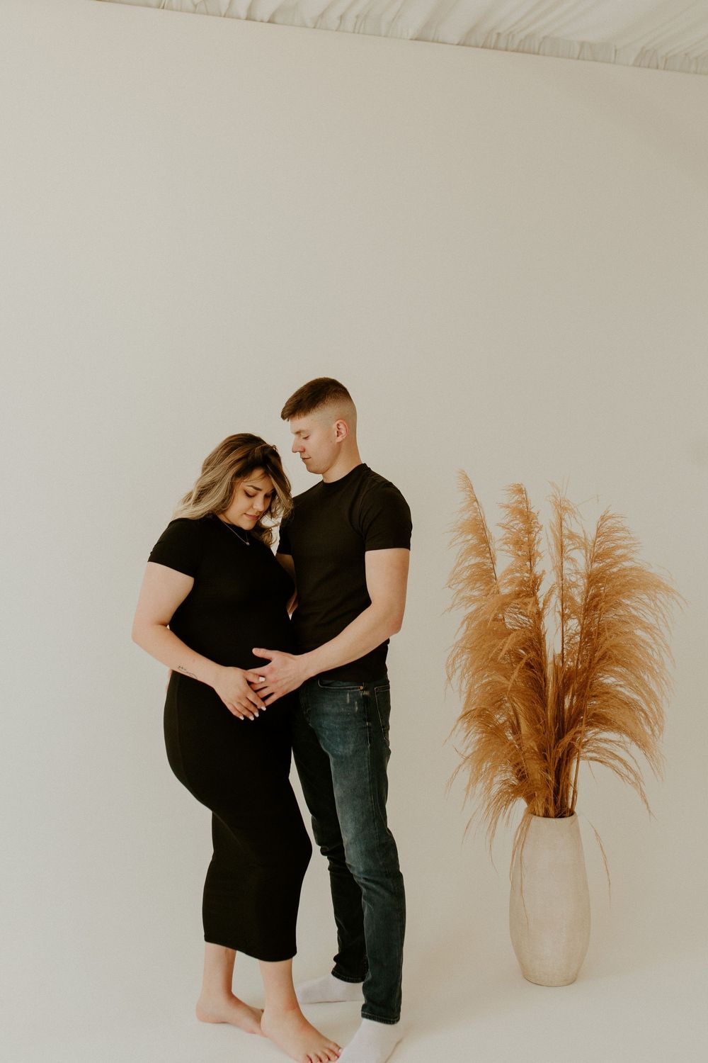 A couple embraces intimately in black outfits next to a vase with pampas grass.