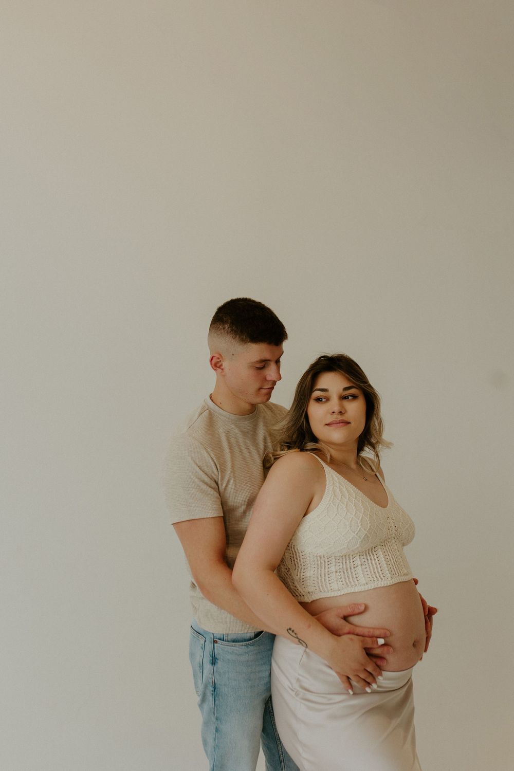Intimate maternity photoshoot featuring couple in casual attire against beige wall.