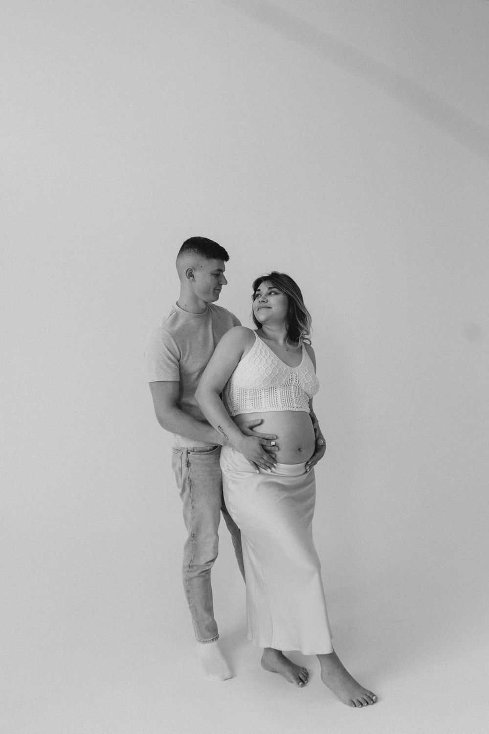 Black and white studio maternity photo of a couple embracing against a white background in minimalist style.