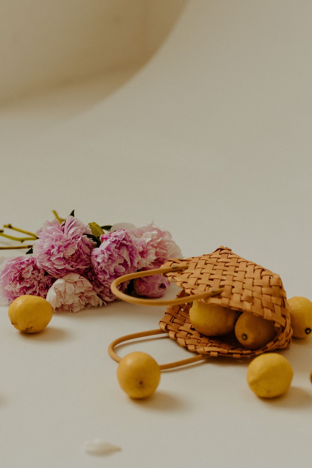Pink peonies and yellow pears arranged artistically with a woven basket against a neutral background in a minimalist style.