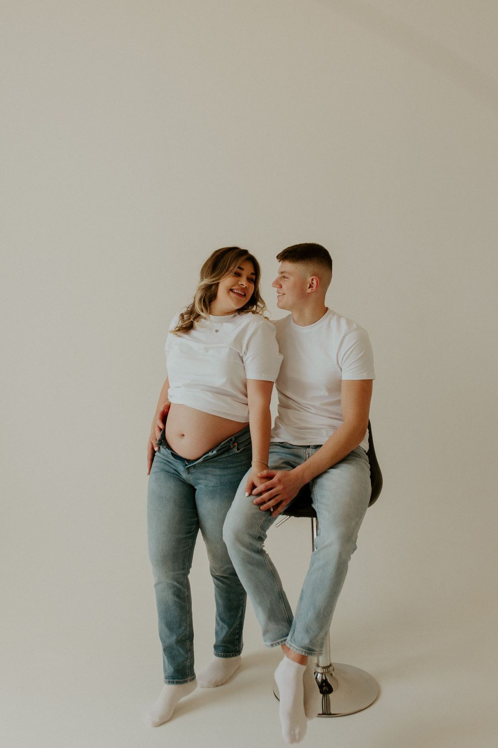 Two people wearing white shirts and jeans sit together against a neutral backdrop in a minimalist studio setting.