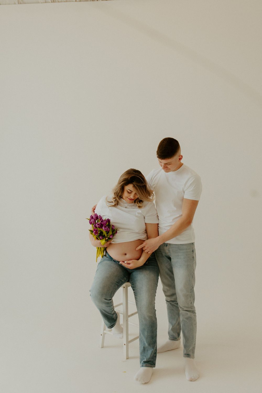 A couple sits together on a white chair holding flowers while embracing a pregnant belly in a minimalist setting.