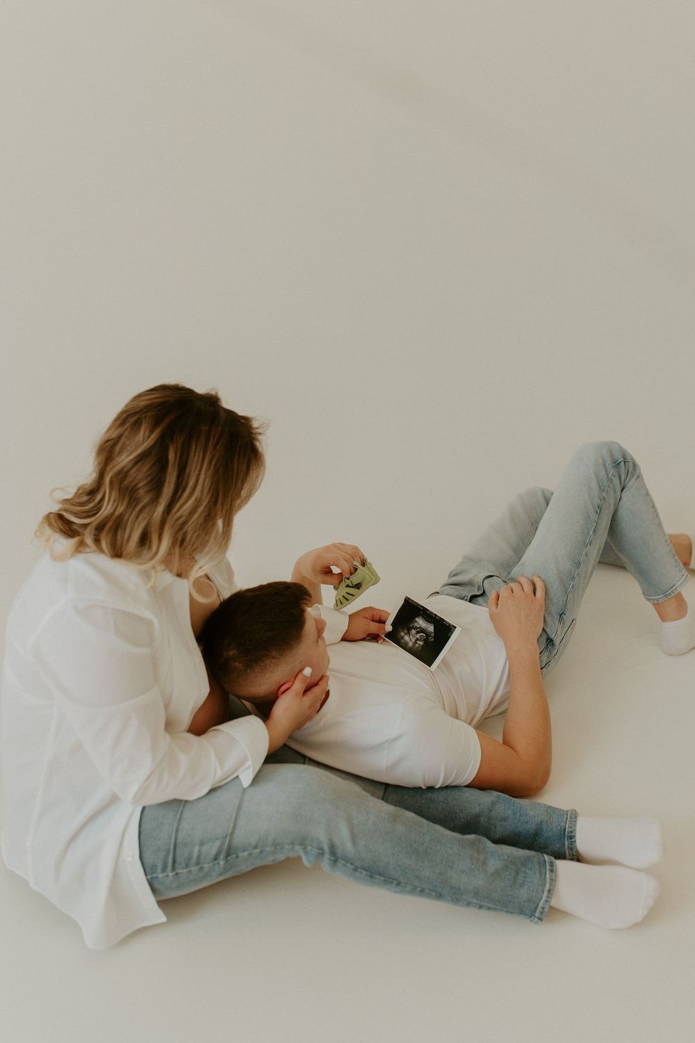 A couple lies together on a white floor looking at an ultrasound photo during a pregnancy photoshoot.