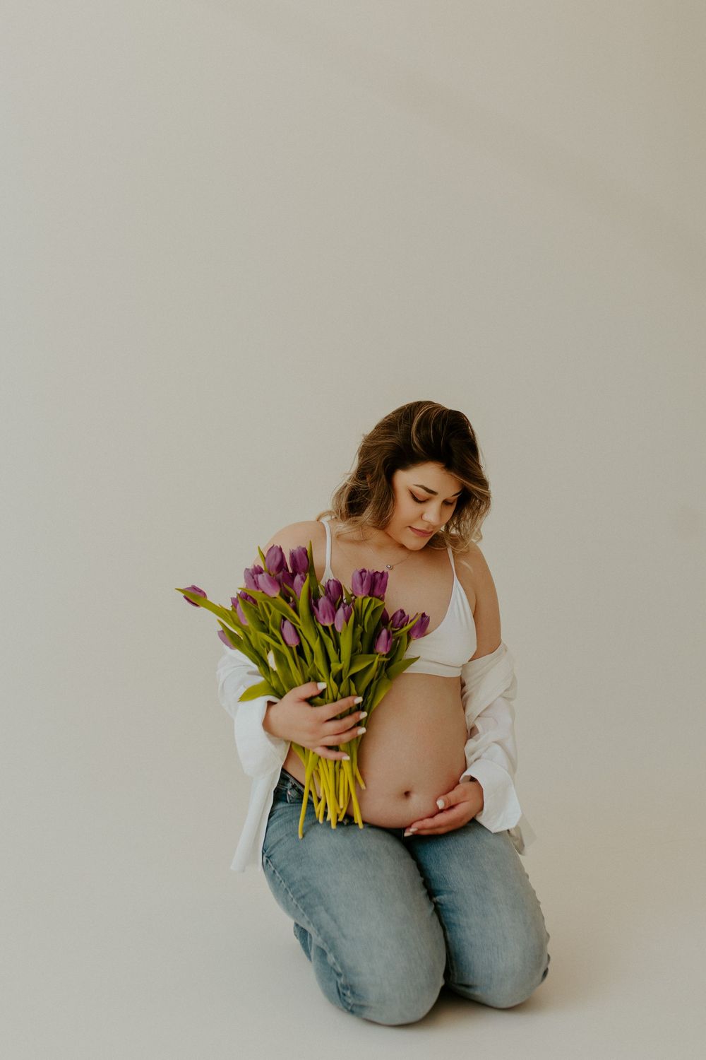Artistic maternity photo series showing pregnant woman in white top holding purple tulips against neutral background.
