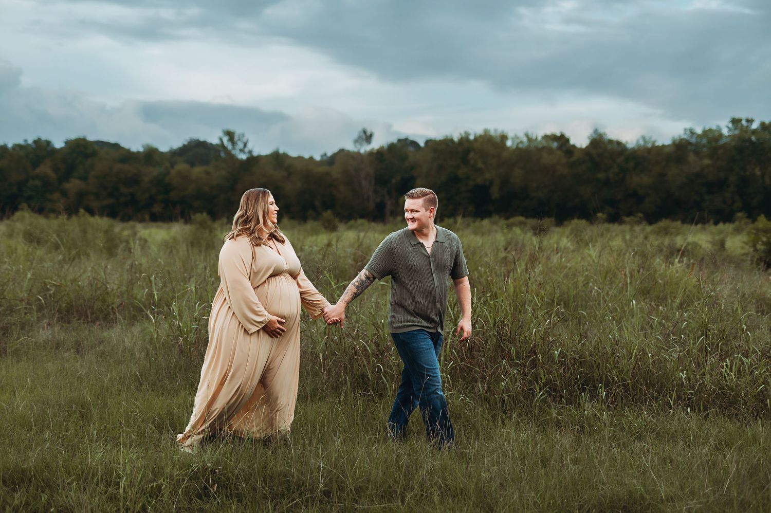 A couple holds hands in a grassy field during a maternity photoshoot with cloudy skies and trees in the background.