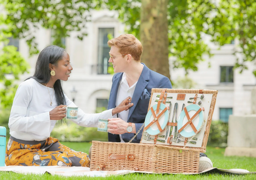 People having a picnic on grass with blanket and basket under green trees.