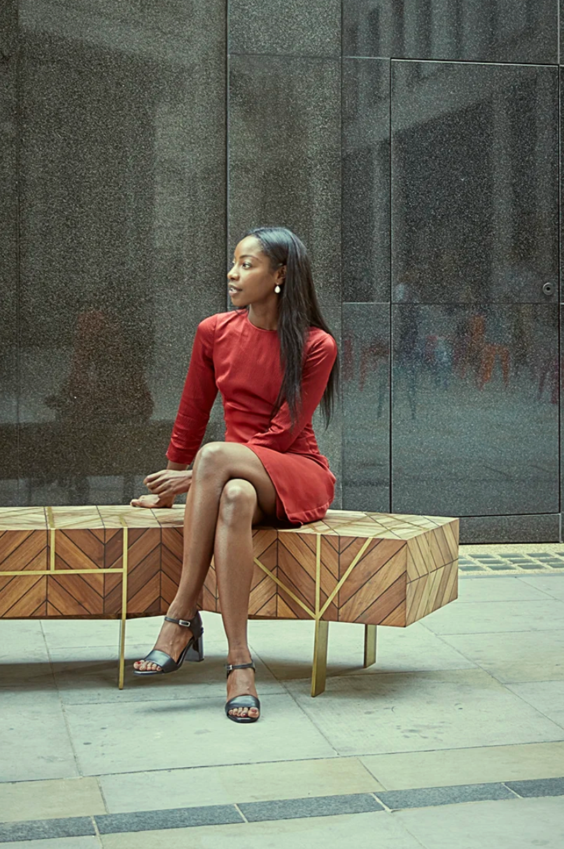 Person in a red dress sitting on a wooden bench outside a modern building.