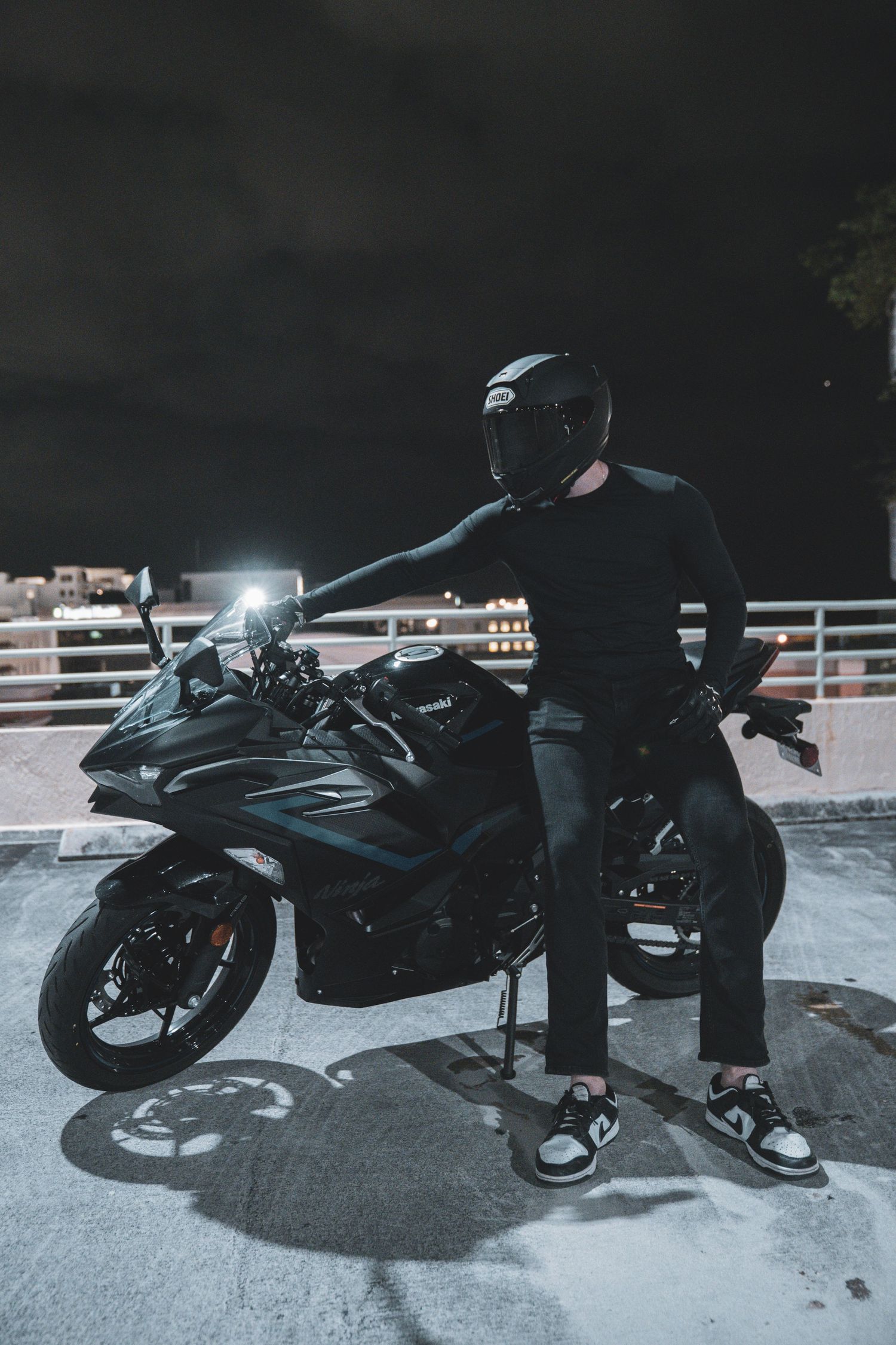 A motorcyclist in dark gear sits on a black sports bike at night, illuminated by ambient lighting.